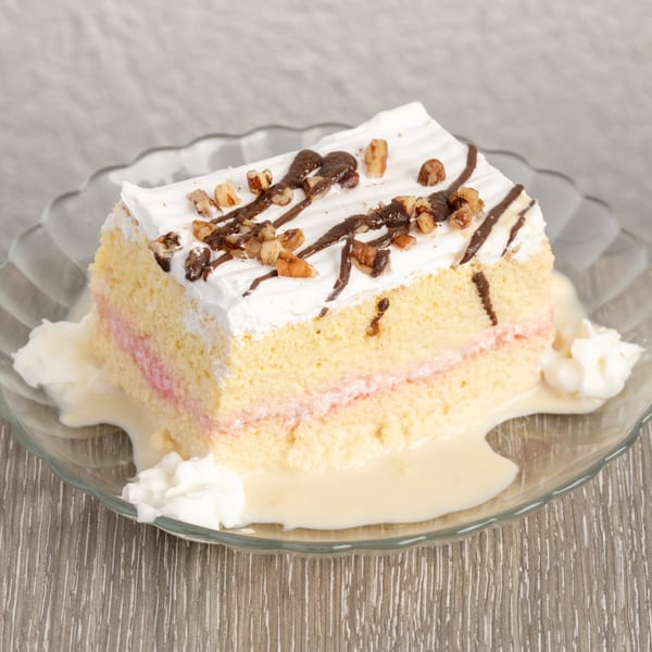 Slice of tres leches cake with whipped cream, chocolate drizzle, and nuts on a glass plate.