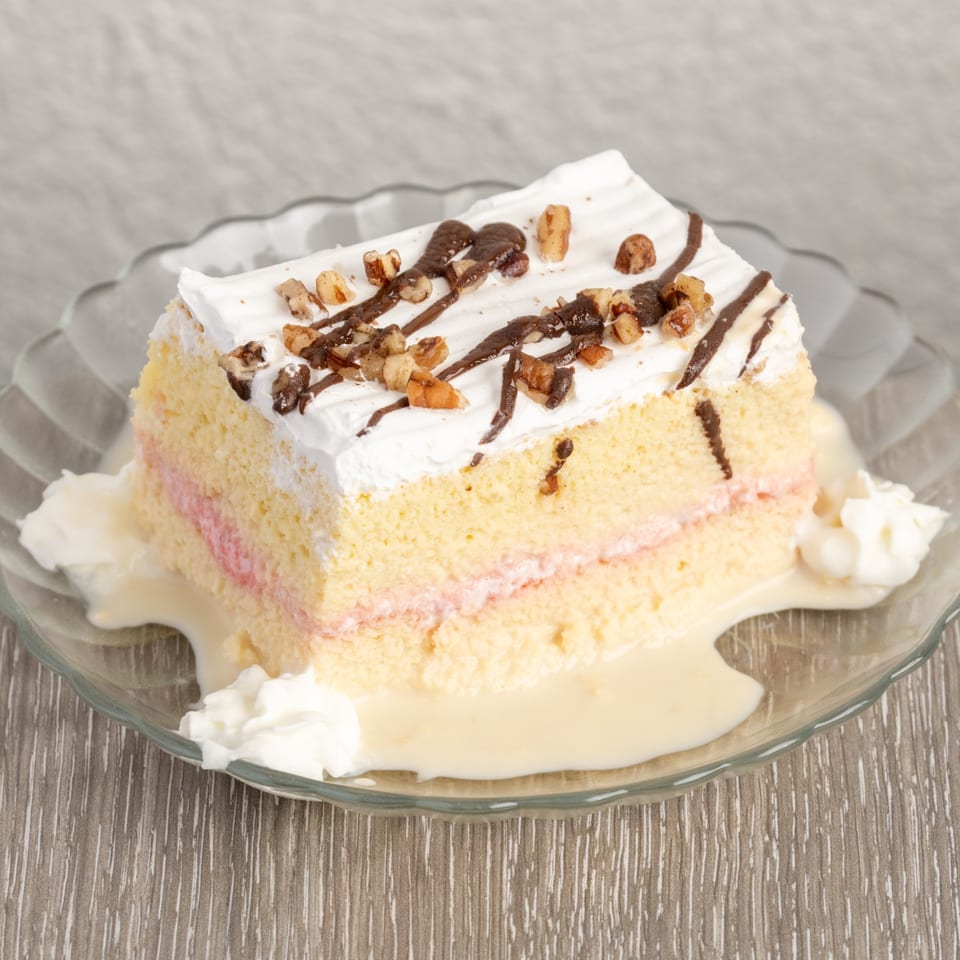 Slice of tres leches cake with whipped cream, chocolate drizzle, and nuts on a glass plate.