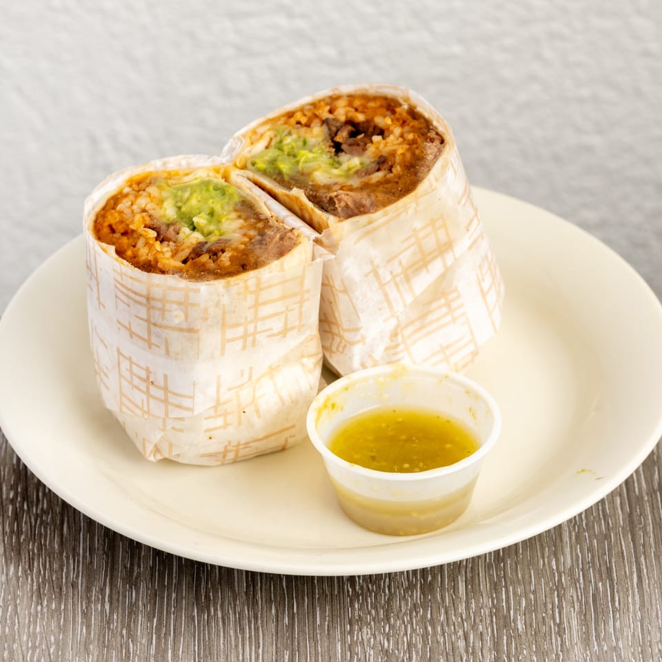 Two burritos, cut in half, revealing rice, beans, and guacamole, with a side of green salsa.