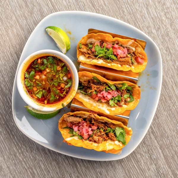 Three birria tacos with cilantro, pickled onions, and a side of consommé with lime wedges.